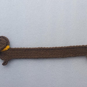 Peut inclure: Un chien teckel au crochet marron fait &agrave; la main avec un collier jaune. Le chien est allong&eacute;, son long corps est &eacute;tir&eacute;. La queue est l&eacute;g&egrave;rement courb&eacute;e. Le chien est fait d'un fil doux et textur&eacute;.