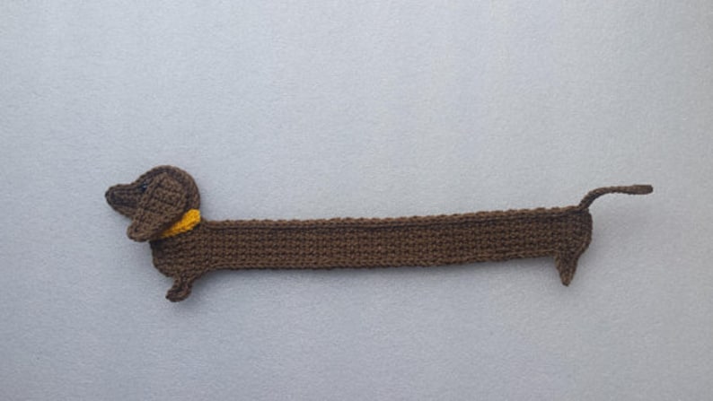 Peut inclure: Un chien teckel au crochet marron fait &agrave; la main avec un collier jaune. Le chien est allong&eacute;, son long corps est &eacute;tir&eacute;. La queue est l&eacute;g&egrave;rement courb&eacute;e. Le chien est fait d'un fil doux et textur&eacute;.