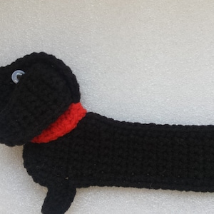 Peut inclure: Un chien teckel noir au crochet avec un collier rouge et des yeux en bouton. Le chien est allong&eacute;, son long corps &eacute;tir&eacute;. Le travail au crochet est d&eacute;taill&eacute; et le chien a une apparence amicale.
