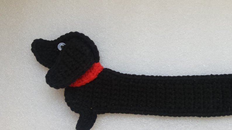 Peut inclure: Un chien teckel noir au crochet avec un collier rouge et des yeux en bouton. Le chien est allong&eacute;, son long corps &eacute;tir&eacute;. Le travail au crochet est d&eacute;taill&eacute; et le chien a une apparence amicale.