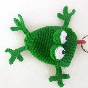 K&ouml;nnte beinhalten: Gr&uuml;ner geh&auml;kelter Frosch-Schl&uuml;sselanh&auml;nger mit wei&szlig;en Augen und einer roten Zunge. Der Frosch liegt auf dem R&uuml;cken mit ausgestreckten Beinen. Der Schl&uuml;sselanh&auml;nger hat einen silbernen Ring.