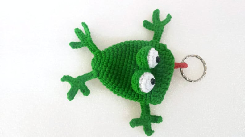 K&ouml;nnte beinhalten: Gr&uuml;ner geh&auml;kelter Frosch-Schl&uuml;sselanh&auml;nger mit wei&szlig;en Augen und einer roten Zunge. Der Frosch liegt auf dem R&uuml;cken mit ausgestreckten Beinen. Der Schl&uuml;sselanh&auml;nger hat einen silbernen Ring.
