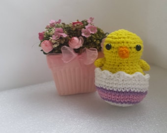 Könnte beinhalten: Ein gelbes gehäkeltes Küken, das aus einem rosa-weiß gestreiften Ei schaut, neben einem rosa Blumentopf mit rosa Blumen.