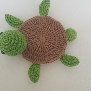 May include: A handmade crochet turtle in shades of brown and green. The turtle has a round, textured brown shell, a green head, and four green flipper-like appendages. Black bead eyes are visible on the head. The turtle is lying on a white surface.