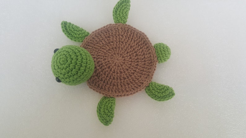May include: A handmade crochet turtle in shades of brown and green. The turtle has a round, textured brown shell, a green head, and four green flipper-like appendages. Black bead eyes are visible on the head. The turtle is lying on a white surface.