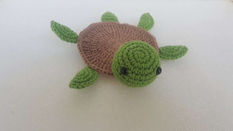 May include: A handmade crochet turtle toy. The turtle has a brown shell, a green head, and green flippers. The eyes are small black beads. The toy is made of soft yarn.
