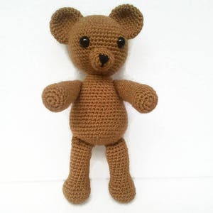 May include: A brown crocheted teddy bear with black eyes. The bear is standing on a white surface.