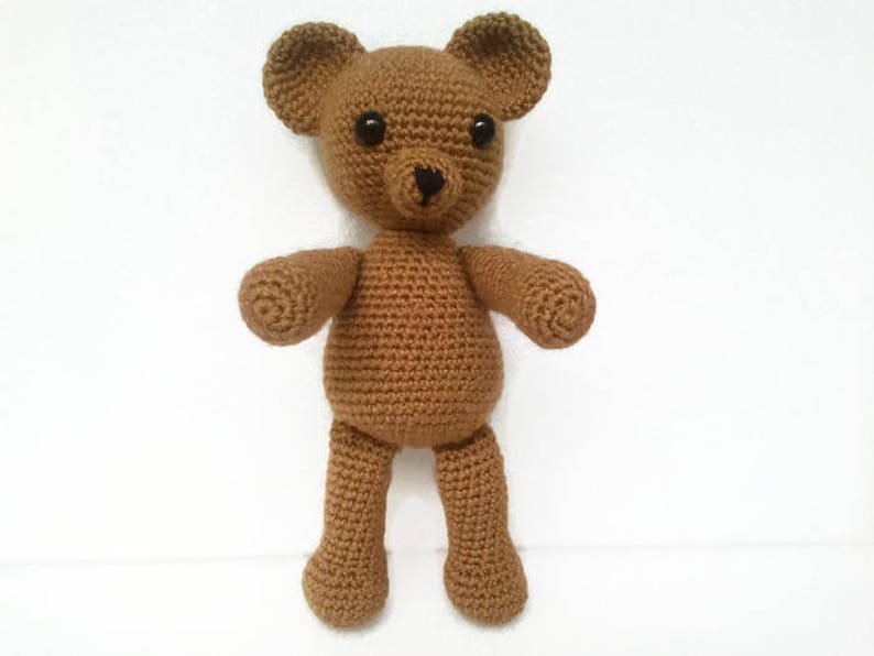 May include: A brown crocheted teddy bear with black eyes. The bear is standing on a white surface.