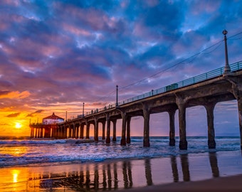 May include: A scenic photograph of a pier extending into the ocean at sunset. The sky is filled with vibrant colors of orange, purple, and blue. The pier's structure is visible, with its pillars reflected in the wet sand.