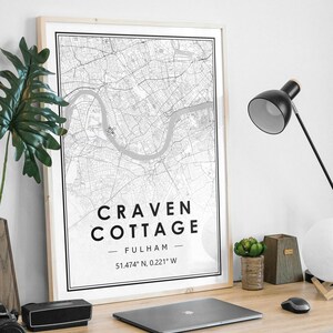 May include: A black and white map print of Craven Cottage in Fulham, London. The print is framed in a light wood frame and is hanging on a white wall. The coordinates 51.474° N, 0.221° W are printed below the name of the location.