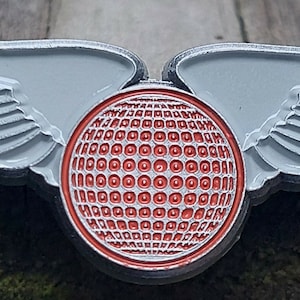 May include: A silver pin with a red and white golf ball design and wings. The wings are silver and have a textured surface.
