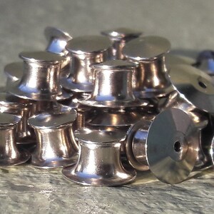 May include: A pile of silver-toned metal safety pin backs. The backs are round and have a small hole in the center.