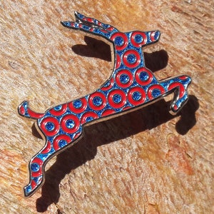 May include: A silver-toned metal pin shaped like a leaping deer. The deer is covered in a pattern of red and blue circles.