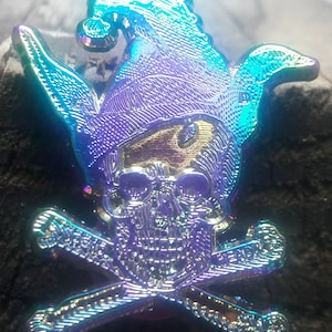 May include: A colorful iridescent metal pin featuring a skull with a jester's hat and crossed bones. The skull and bones are detailed with a textured design.