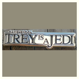 May include: A silver-colored metal plaque with the text "GAMEHENDGE TREY IS A JEDI" in raised lettering. The plaque is rectangular with a slightly curved bottom edge and is set against a wooden background.