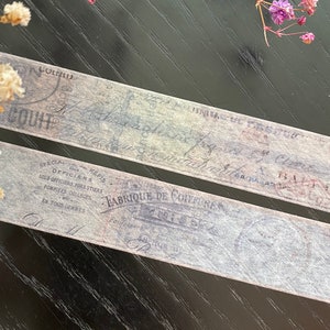 Pode incluir: Duas tiras de fita washi vintage com tons azul e cinza desbotados, com texto e selos franceses. Delicadas flores secas em tons de branco e roxo são dispostas ao redor da fita em uma superfície de madeira escura.