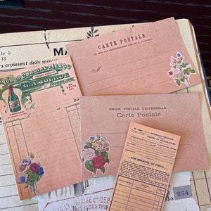 May include: A collection of vintage-style paper ephemera, including postcards and receipts, in shades of peach and cream. Floral stickers adorn the items, with text in French. The items are arranged on a dark surface.