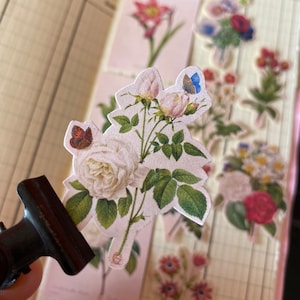 May include: A set of vintage floral stickers featuring various flowers and butterflies. The stickers are printed on a white background and have a textured finish. The flowers include roses, lilies, and daisies. The butterflies are in shades of blue, red, and white.