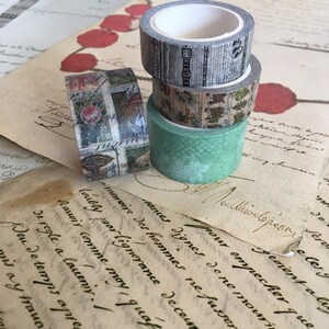 May include: Four rolls of decorative washi tape in various designs. One roll features a newspaper print, another has a floral pattern, and a third has a green textured design. The fourth roll has a rose and text design. The rolls are stacked on top of each other.