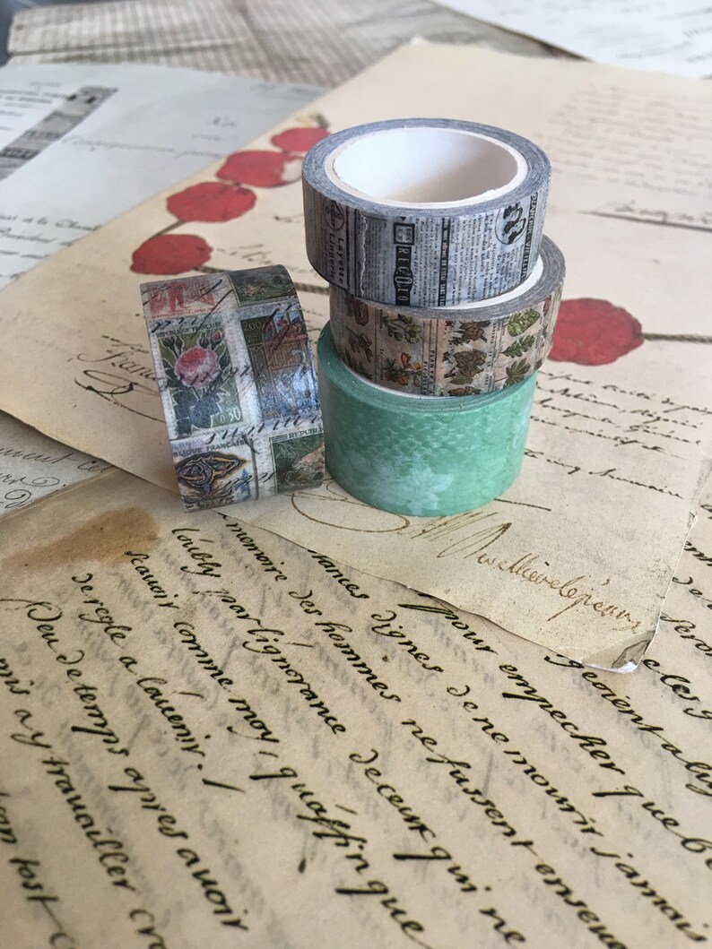 May include: Four rolls of decorative washi tape in various designs. One roll features a newspaper print, another has a floral pattern, and a third has a green textured design. The fourth roll has a rose and text design. The rolls are stacked on top of each other.