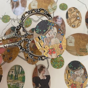 May include: Silver scissors holding an oval sticker with a detail from Gustav Klimt's "The Kiss." Other oval stickers with similar artwork and images of women are scattered around. The background is a map.