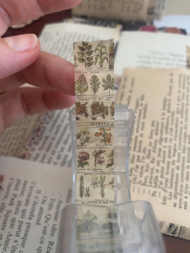 May include: A roll of botanical-themed washi tape featuring illustrations of leaves and flowers. The tape is held in a clear dispenser, with pages of a book in the background. The tape has a vintage, antique aesthetic.