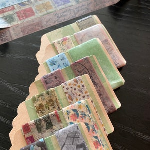 May include: A set of eight rolls of decorative washi tape with various patterns, including floral, vintage, and geometric designs. The tape is packaged on cardboard spools with a green and beige colour scheme.