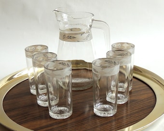 May include: A clear glass pitcher and six matching glasses with a decorative band featuring a leaf pattern. The glasses and pitcher are arranged on a round wooden tray with a gold rim. The pitcher has a handle.