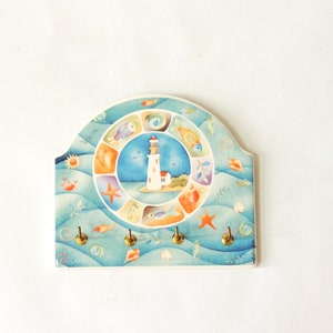 May include: A ceramic key holder shaped like a half-circle with a lighthouse and sea life design. The key holder has five hooks and is painted in shades of blue, green, orange, and yellow.