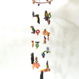May include: A colorful, handcrafted mobile featuring various wooden bird and animal figures. The mobile includes birds in green, yellow, and black, along with other abstract shapes and figures. The figures are suspended from a wooden ring at the top.