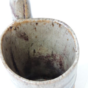 K&ouml;nnte beinhalten: Eine Keramiktasse mit einem Henkel in Form eines Ausgusses. Die Tasse ist in einem hellgrauen Ton glasiert mit braunen Flecken und Streifen.