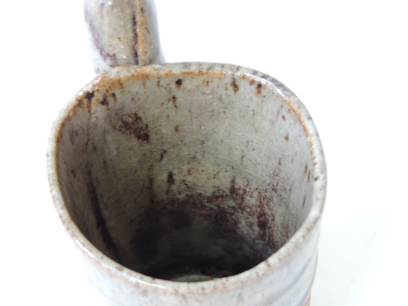 K&ouml;nnte beinhalten: Eine Keramiktasse mit einem Henkel in Form eines Ausgusses. Die Tasse ist in einem hellgrauen Ton glasiert mit braunen Flecken und Streifen.