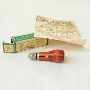 Op de afbeelding: Een vintage naald met een houten handvat en een metalen punt, in de originele doos met instructies. De doos is groen en wit met de tekst "The Speedy Stitcher".