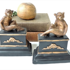 May include: A pair of gold monkey bookends with black bases. Each monkey is sitting on a black base with gold accents. The bases have a gold trim around the top and bottom.