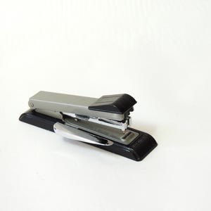May include: A silver and black stapler on a white background. The stapler has a black base and top, with a silver metal stapling mechanism and a chrome lever. Office supply.
