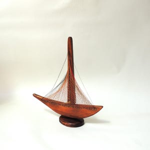 Peut inclure: Maquette de voilier en bois. La coque est marron foncé, avec un mât élancé. Les voiles sont représentées par un réseau de fils fins colorés en rouge, noir et orange. Le modèle repose sur un socle rond en bois, pour la décoration.