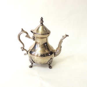 Może przedstawiać: Srebrny czajnik z ozdobnym uchwytem i dzióbkiem. Czajnik ma okrągły korpus i stoi na trzech nóżkach.