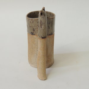 K&ouml;nnte beinhalten: Eine Keramiktasse mit einem einzigartigen, handgefertigten Henkel. Die Tasse ist in einem gesprenkelten Braun und Beige glasiert.