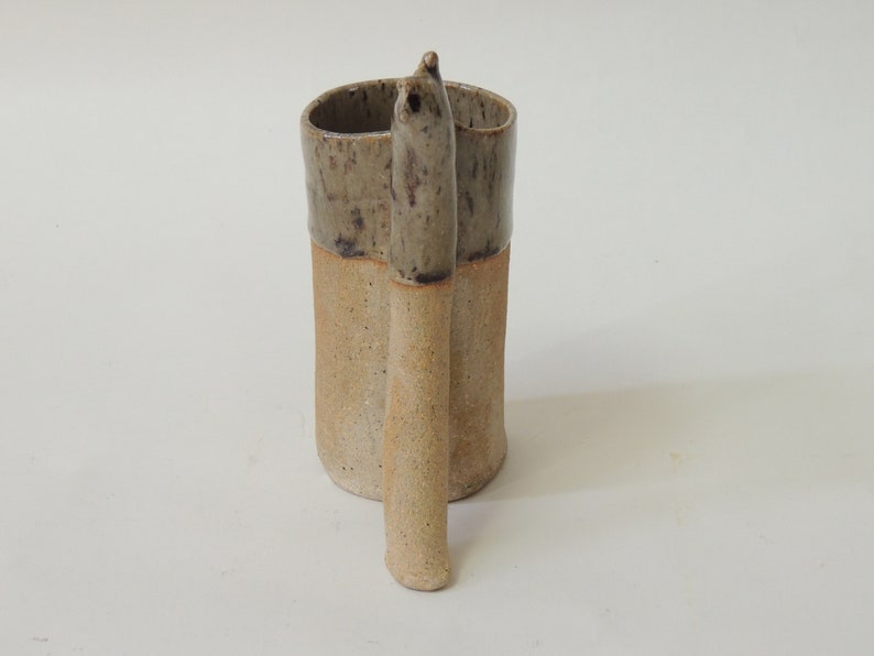 K&ouml;nnte beinhalten: Eine Keramiktasse mit einem einzigartigen, handgefertigten Henkel. Die Tasse ist in einem gesprenkelten Braun und Beige glasiert.