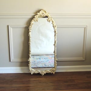 May include: An ornate, ivory-colored, full-length mirror with a decorative frame. The mirror has a tall, arched top and a detailed, floral-like design around the edges. It leans against a neutral-colored wall and sits on a wooden floor.