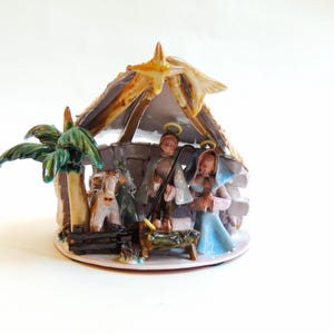 May include: A ceramic nativity scene featuring a stable with a star, palm tree, and figures of Mary, Joseph, and baby Jesus. The figures are in muted colors, with a cow and donkey. The scene is on a round base.