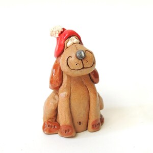 May include: A ceramic dog figurine with a red hat and a white pom-pom. The dog has a smiling face, brown fur, and a silver nose. The figurine is sitting down.
