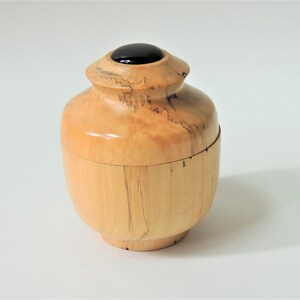 May include: A small wooden box with a lid, made of light wood with dark brown streaks. The lid has a black circular inlay in the center.