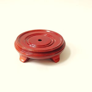 May include: A round, reddish-brown wooden display stand with a smooth, polished finish. The stand features a raised circular platform with a small, centered hole and is supported by three short, curved feet that elevate it from the surface.