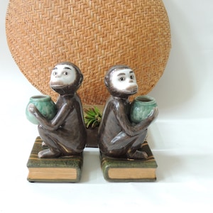 May include: A pair of bookends featuring brown ceramic monkeys holding green ceramic pots. The monkeys are sitting on top of books with green and brown covers.