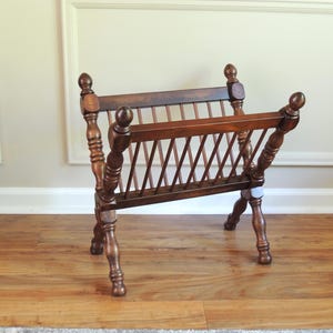 May include: A wooden magazine rack with a slatted design and turned legs. The rack is made of dark brown wood and has a rustic look.