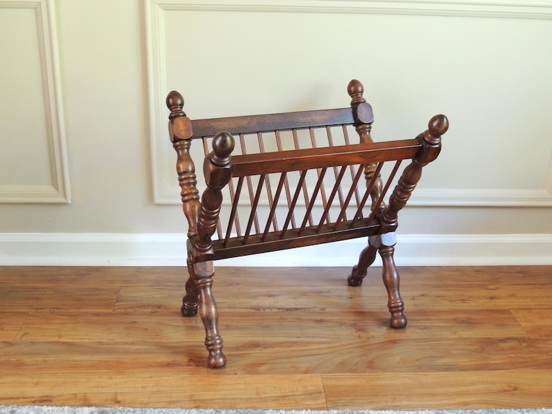 May include: A wooden magazine rack with a slatted design and turned legs. The rack is made of dark brown wood and has a rustic look.