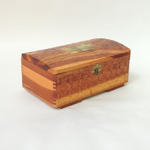 May include: A wooden box with a curved lid and floral pattern. The box is made of light and dark brown wood, with a brass clasp. The lid has a rectangular decorative element. The box is rectangular and sits on small feet.