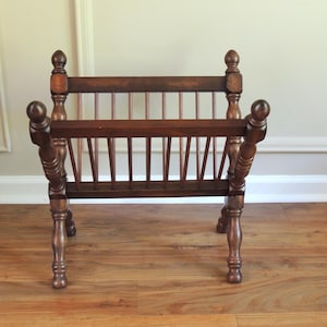 May include: A wooden magazine rack with a slatted design and turned legs. The rack is made of dark brown wood and has a rustic look.