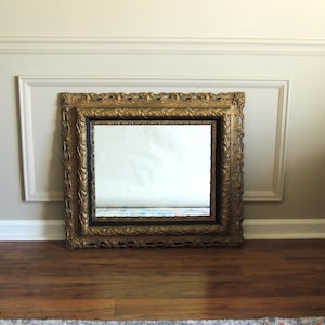 May include: A rectangular mirror with an ornate, gold-colored frame. The frame features detailed carvings and a dark inner border. The mirror is resting on a wooden floor against a light-colored wall with decorative molding.
